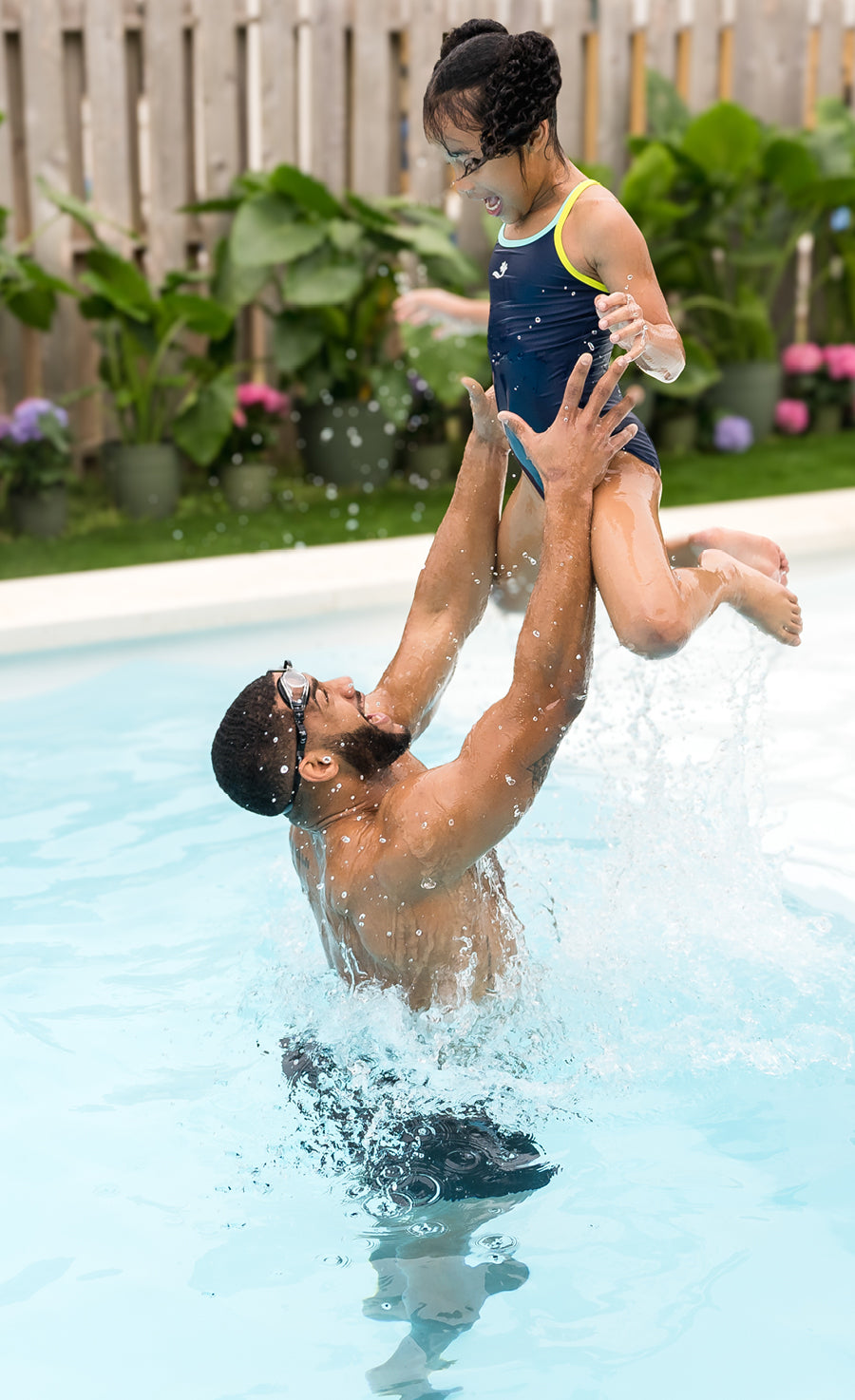 The image captures a joyful moment in a swimming pool. A man and a young girl are engaged in playful activity. The man, wearing goggles and no shirt, is partially submerged in the water and is lifting the girl up above the surface. The girl, in a blue swimsuit with yellow trim, is laughing and appears to be having a great time as she is tossed into the air by the man. The setting is outdoors with a visible fence and lush greenery in the background, indicating a private or residential pool area. The interact
