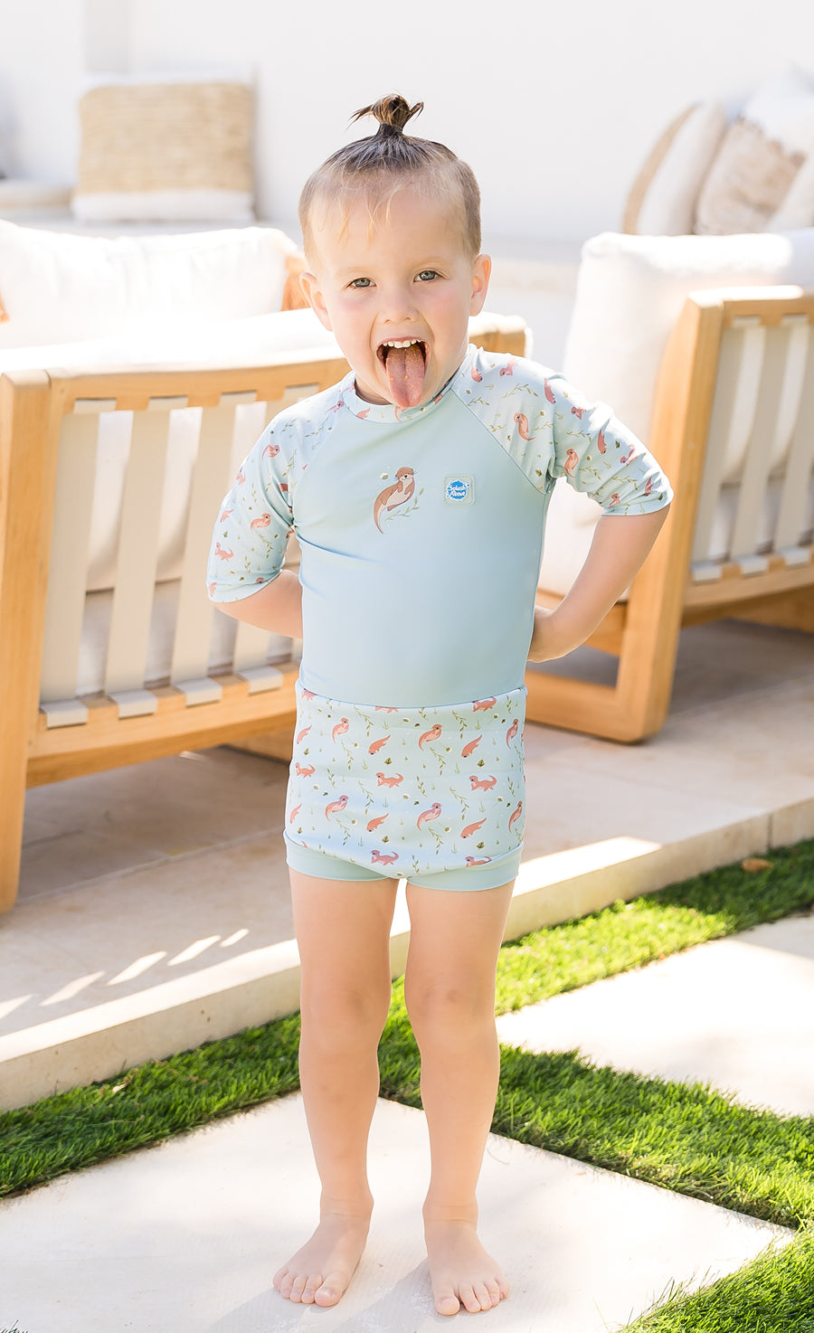 The image features a young toddler standing on a patio with a joyful expression, sticking out their tongue. The child is wearing a two-piece swimwear set with a light blue top and shorts adorned with a pattern of small, colorful sea creatures. The patio is bordered by a well-maintained lawn and furnished with wooden outdoor chairs. The setting suggests a warm, sunny day perfect for outdoor activities. This photo captures the essence of childhood exuberance and the joy of summertime play.