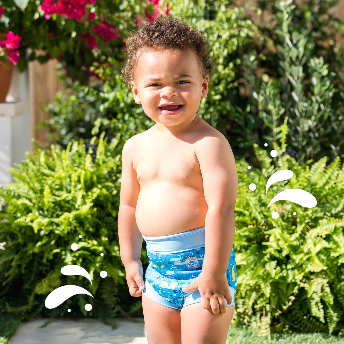 The image shows a young child standing outdoors on a sunny day, wearing a blue swim diaper with a playful sea creature design. The child has a joyful expression and curly hair. The background features lush greenery and pink flowers, indicating a well-maintained garden or park. The scene captures the essence of summer fun and the innocence of childhood. This photo could be used to illustrate concepts related to summer activities, children's swimwear, or outdoor leisure for young children.