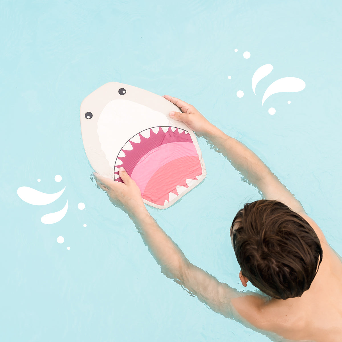The image depicts a playful scene in a swimming pool where a person is holding an inflatable shark toy above the water. The shark has a cartoonish design with a white body, a large pink mouth with visible teeth, and two black eyes. The person is only partially visible from the back, with their arms raised, holding the shark. The water is a light blue color, and there are white bubbles around the shark, suggesting movement or recent activity. The scene captures a moment of leisure and fun, likely intended fo